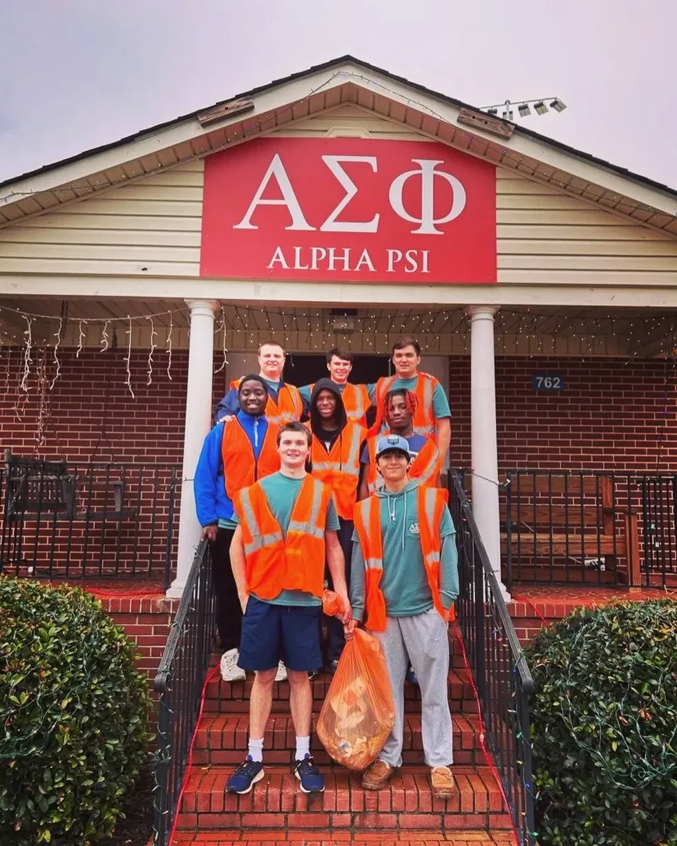 Brothers of Alpha Psi in safety vests holding trash bags on the front steps of the chapter house during a community service project